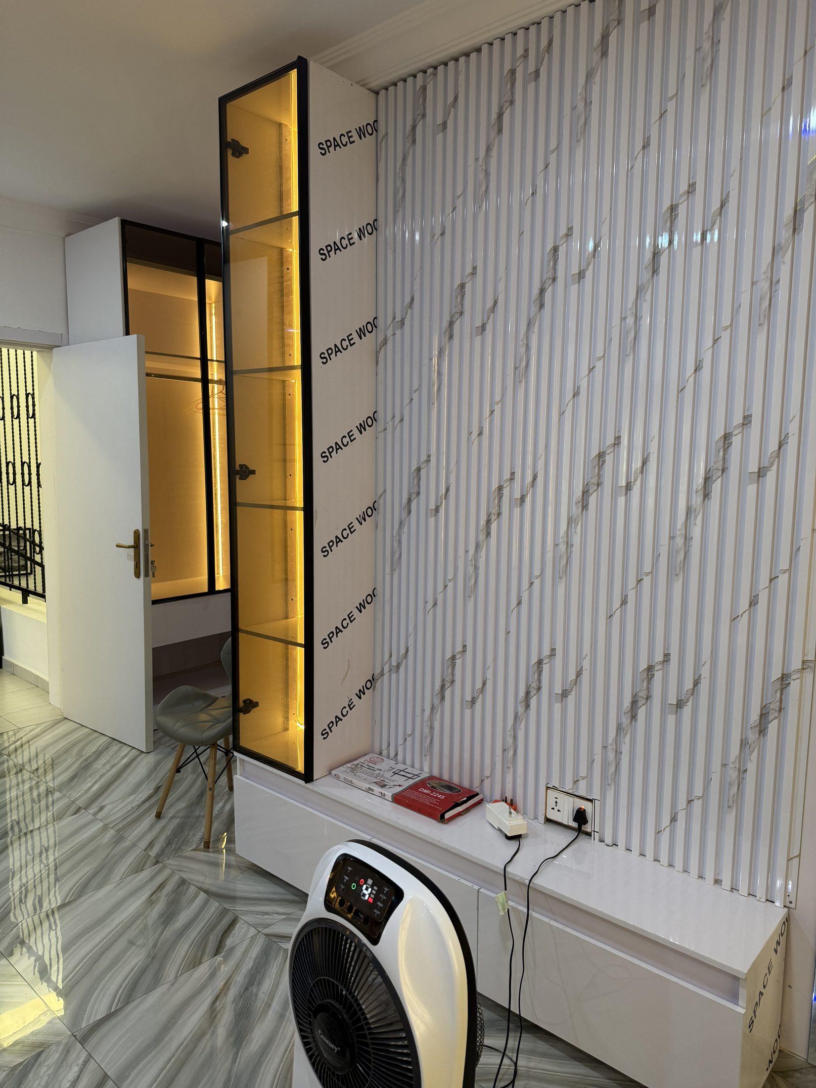 Tall black-framed glass display cabinet with warm shelf lighting beside a marble-effect fluted wall panel, photographed during installation