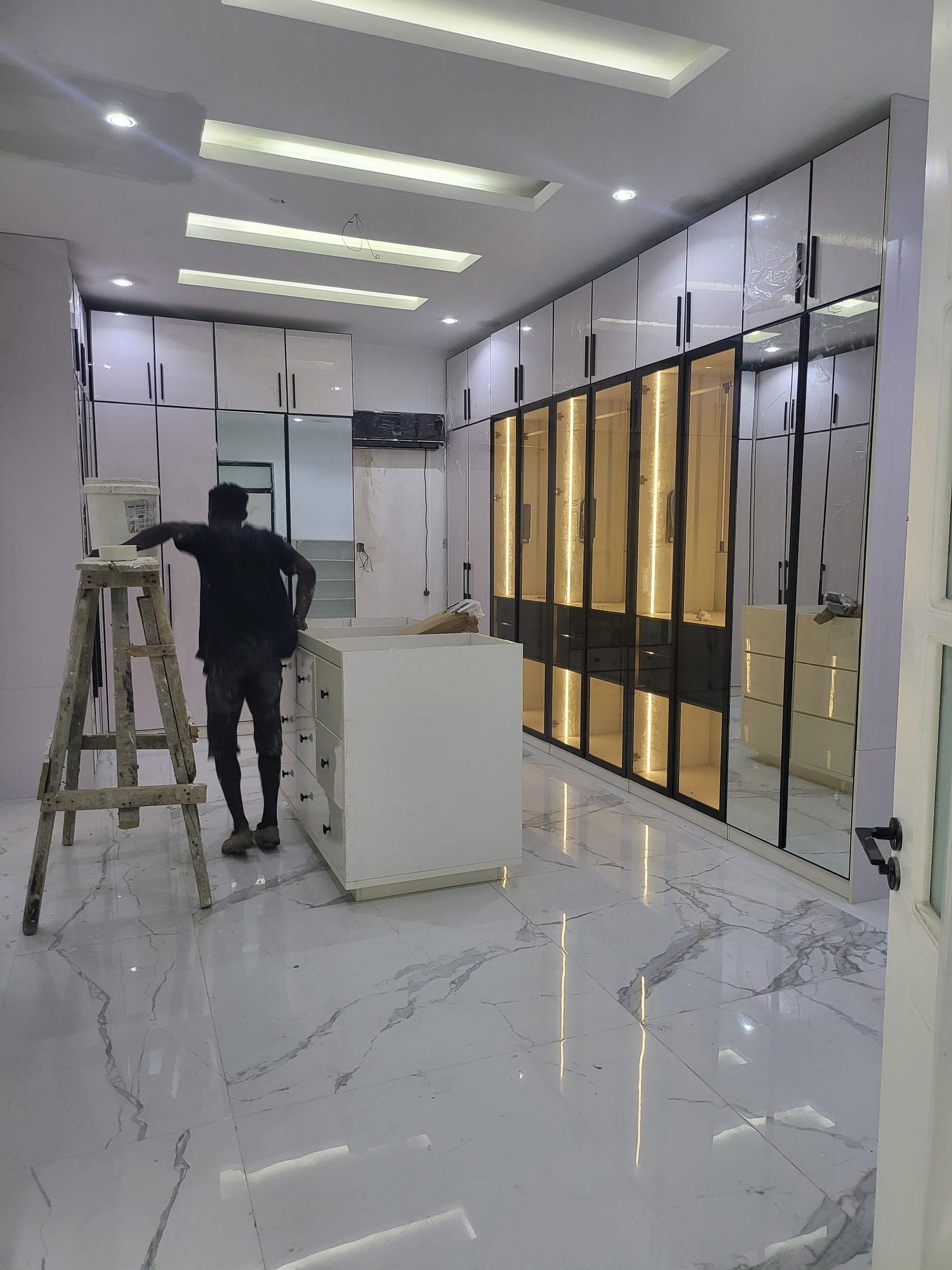 Dressing room with lit black-framed glass wardrobe doors, pale gloss cabinets and a marble-effect floor, with a fitter at work
