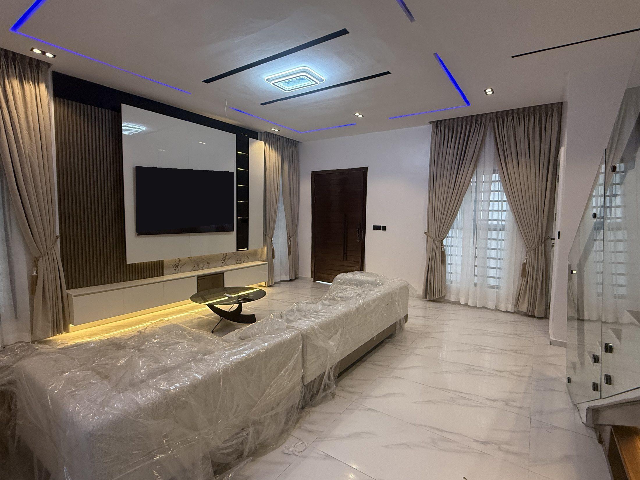 Wide view of a living room with a gloss media wall, blue-lit ceiling details, full-height curtains and a glass staircase