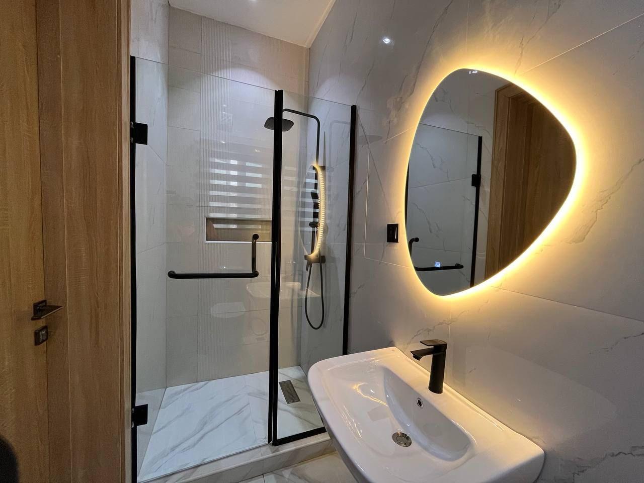 Marble-tiled bathroom with a glowing organic mirror, black shower fittings and a glass enclosure