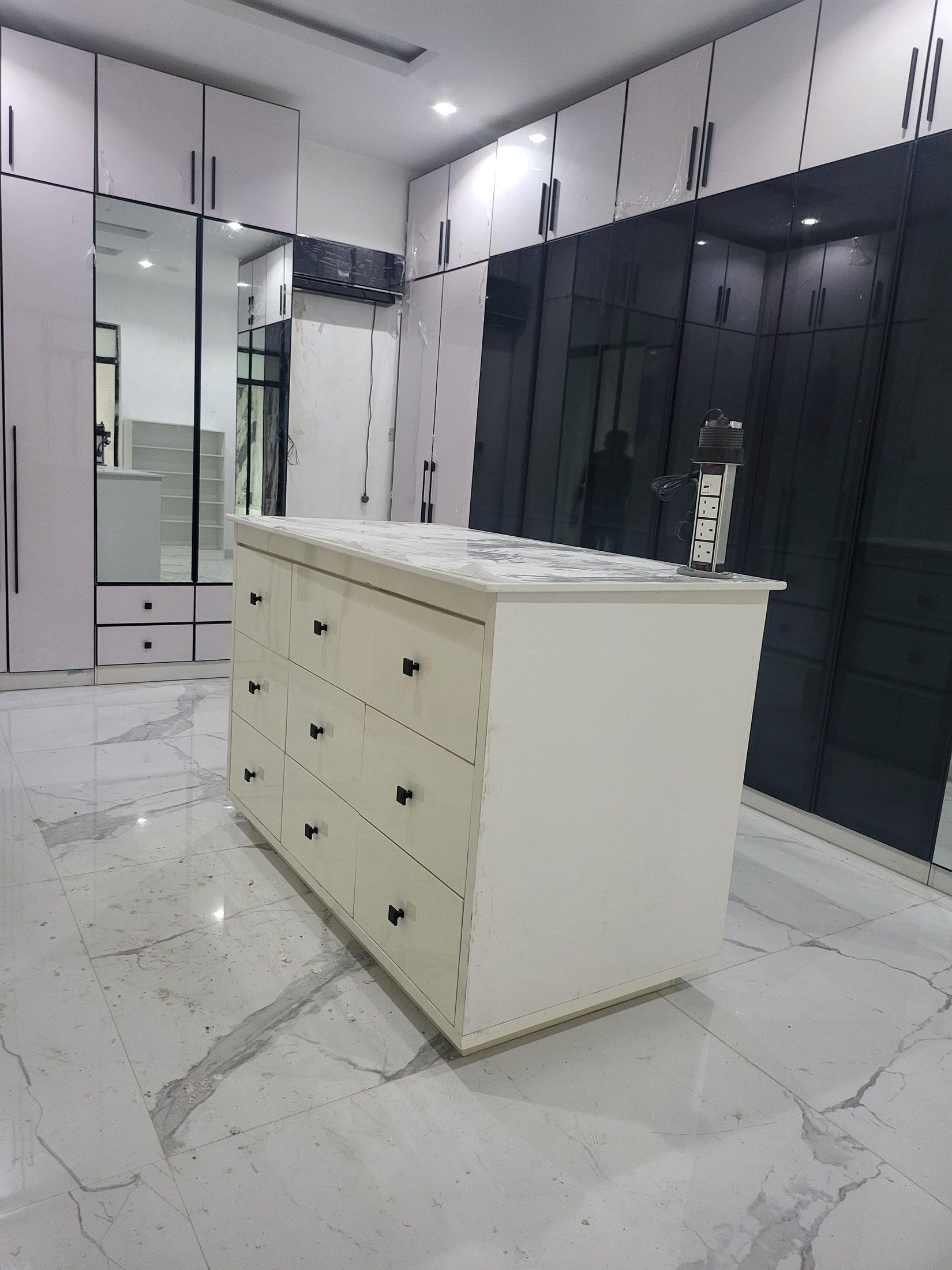 Dressing room island with cream drawers and black knobs, set between pale gloss wardrobes, full-length mirrors and black glass doors