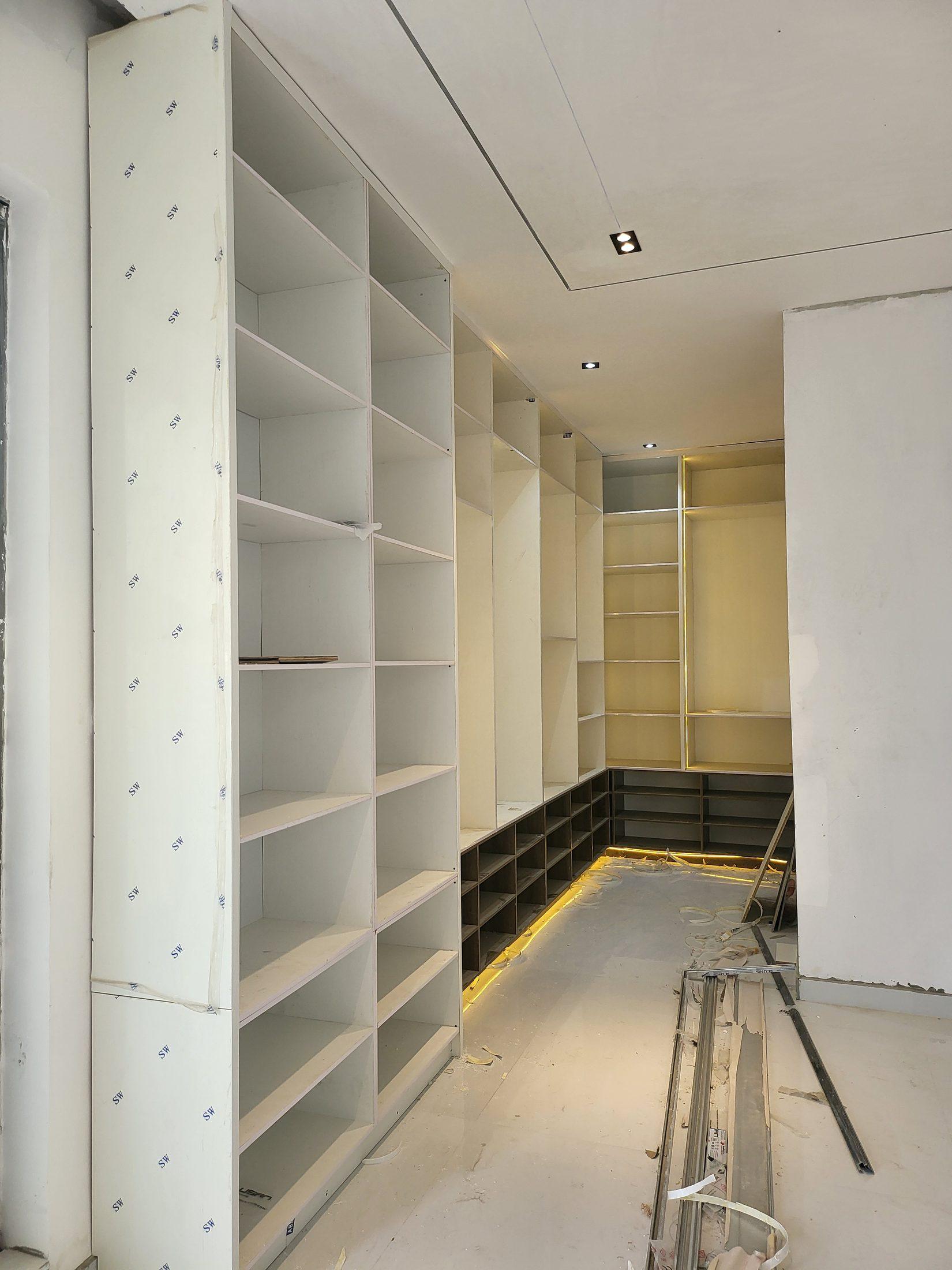 White walk-in wardrobe with full-height shelving, walnut shoe cubbies and warm plinth lighting